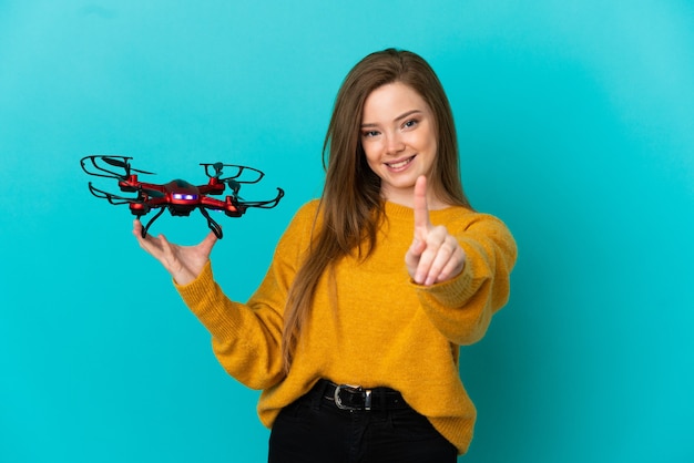 Tienermeisje dat een drone houdt over een geïsoleerde blauwe achtergrond die een vinger laat zien en optilt