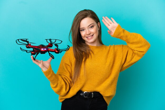 Tienermeisje dat een drone over geïsoleerde blauwe achtergrond houdt en met de hand salueert met een gelukkige uitdrukking