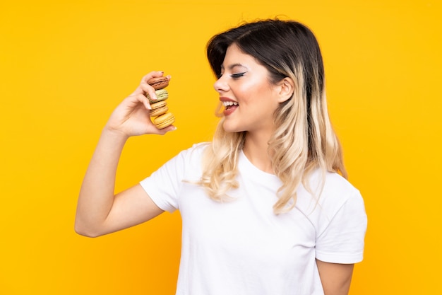 Tienermeisje dat op gele muur wordt geïsoleerd die kleurrijke Franse macarons houdt en een overwinning viert