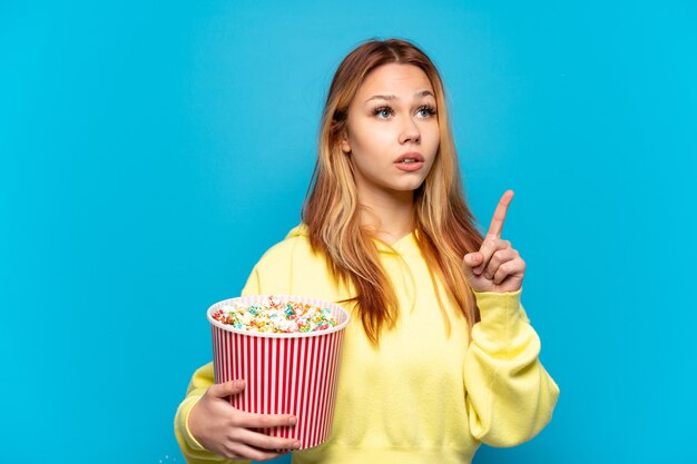 Tienermeisje dat popcorns vasthoudt over geïsoleerde blauwe achtergrond en denkt aan een idee dat de vinger omhoog wijst