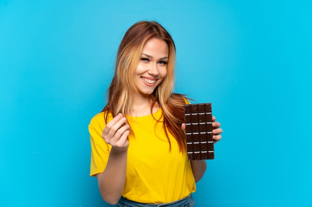 Tienermeisje die chocolade houden over geïsoleerde blauwe achtergrond die geldgebaar maakt