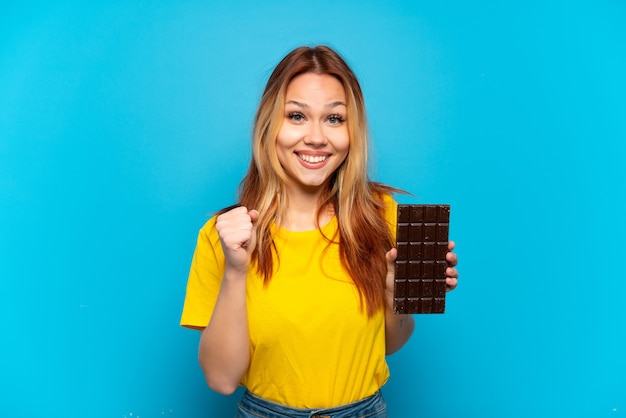 Tienermeisje die chocolade over geïsoleerde blauwe achtergrond houden die een overwinning in winnaarpositie viert