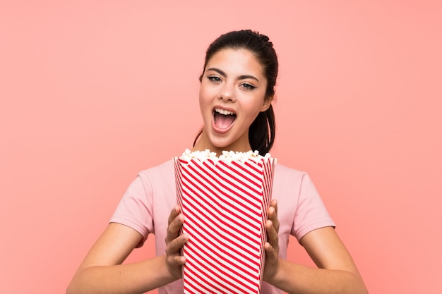 Tienermeisje die over geïsoleerde roze muur popcorns eten die verrassingsgebaar doen