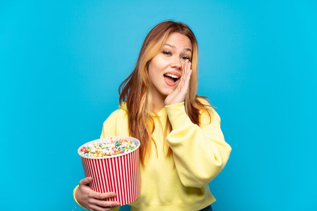 Tienermeisje die popcorns over geïsoleerde blauwe achtergrond houden die met wijd open mond schreeuwen