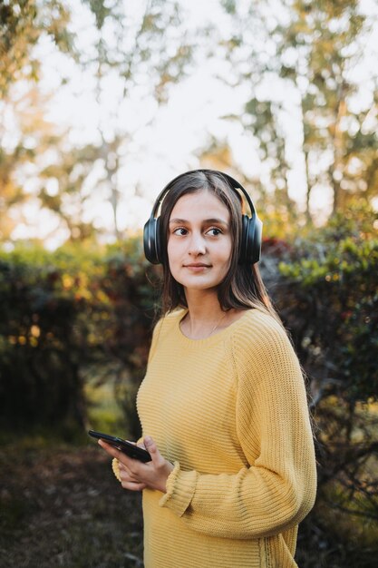 Tienermeisje draagt gele trui, met koptelefoon voor het afspelen van muziek op haar smartphone buiten.