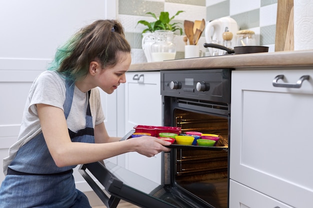 Tienermeisje in een schort met het voorbereiden van rauwe cupcakes in siliconen mallen, dienblad in de oven, achtergrond interieur van thuis koken. Hobby's voor tieners, thuis bakken, zelfgemaakte gezonde voeding