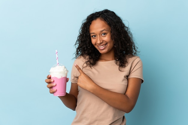 Tienermeisje met aardbeimilkshake die op blauw wordt geïsoleerd dat naar de kant wijst om een product te presenteren