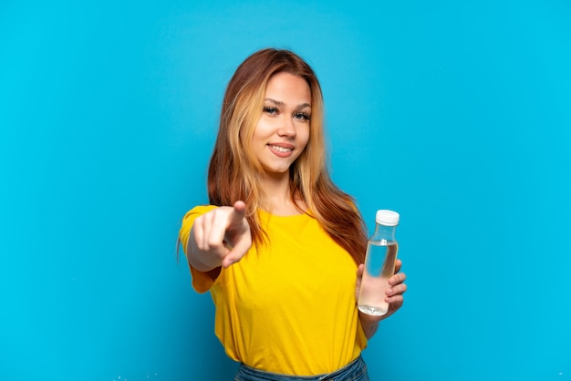 Tienermeisje met een fles water over geïsoleerde blauwe achtergrond die naar voren wijst met gelukkige uitdrukking