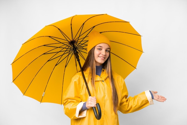 Tienermeisje met regenbestendige jas en paraplu over geïsoleerde witte achtergrond die handen naar de zijkant uitstrekt om uit te nodigen om te komen