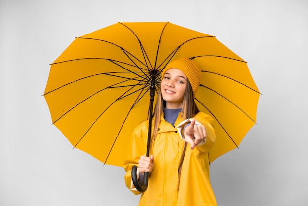 Tienermeisje met regenbestendige jas en paraplu over geïsoleerde witte achtergrond wijzend naar voren met gelukkige uitdrukking