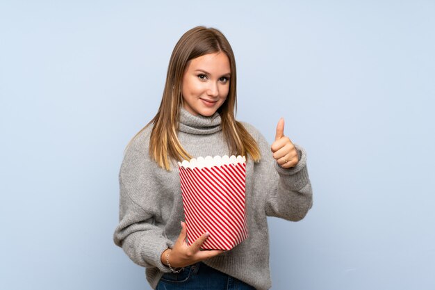 Tienermeisje met sweater over geïsoleerde blauwe muur die een kom popcorns houden