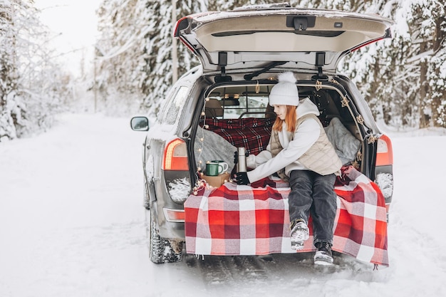 Tienermeisje met thermos zit in de kofferbak van de auto versierd voor Kerstmis Road trip avontuur en lokaal reisconcept Winterpicknick in besneeuwd bos Nieuwjaarsvakantie