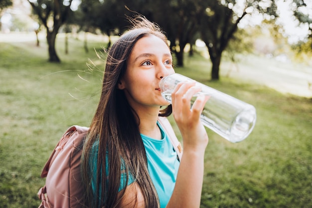 Tienermeisje met turquoise t-shirt en roze rugzak, drinkwater uit glazen fles in het park