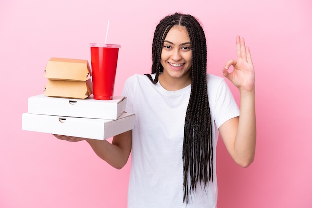 Tienermeisje met vlechten die pizza's en hamburgers houden over geïsoleerde roze achtergrond die ok teken met vingers toont