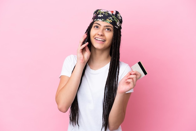 Tienermeisje met vlechten over geïsoleerde roze achtergrond die een gesprek voert met de mobiele telefoon en een creditcard vasthoudt