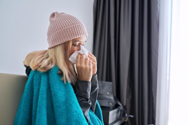 Tienermeisje niezen in zakdoek, vrouw met ziektesymptomen om thuis te zitten bedekt met warme deken en gebreide muts te dragen