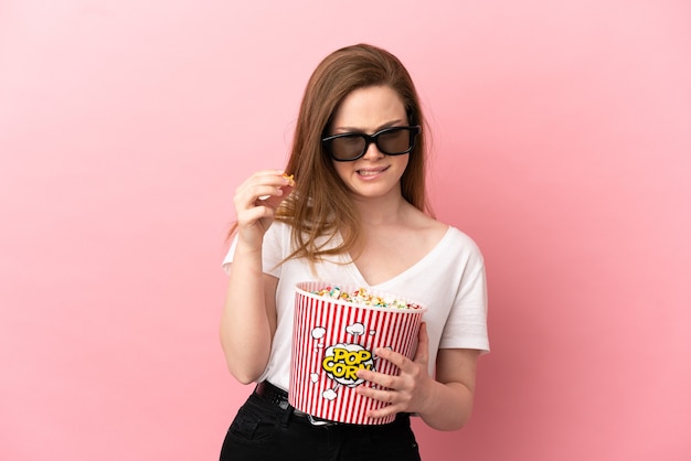 Tienermeisje over geïsoleerde roze achtergrond met 3D-bril en met een grote emmer popcorns