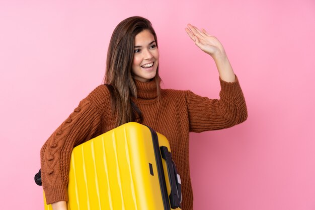 Tienermeisje over geïsoleerde roze muur in vakantie met reiskoffer en het groeten