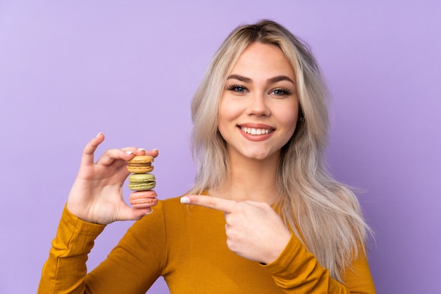 Tienermeisje over purpere muur die kleurrijke Franse macarons houden en het richten