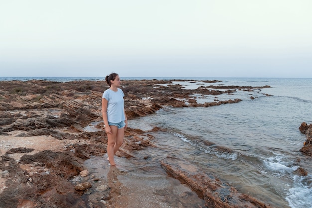 Tienermeisje staande op een klif aan zee bij zonsondergang met een lichtblauw t-shirt, een spijkerbroek en wegkijkend. T-shirtmodel