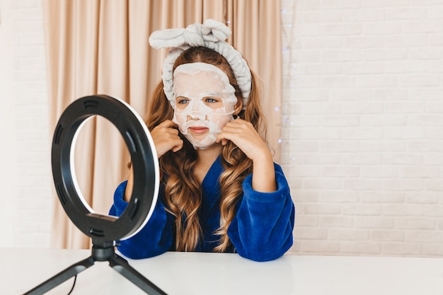 Tienermeisje vlogger die gezichtsmasker voor huidverzorging toepast tijdens het opnemen van video voor een lichtlamp. Gelukkig meisje met plezier bij het filmen van tutorial met behulp van mobiele smartphone.