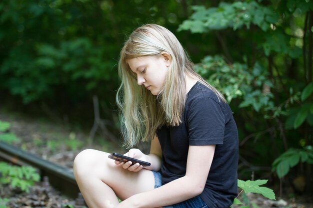 Tienermeisje zittend op het spoor in het bos met smartphone op zomerdag