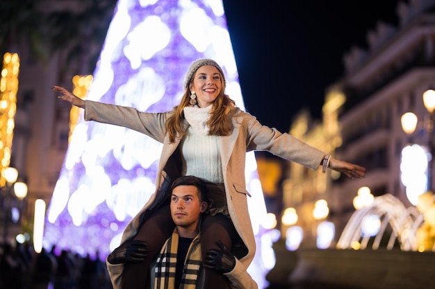 Tienerpaar met kerstnacht plezier buitenshuis. guy draagt meisje op zijn schouders met uitgestrekte armen.