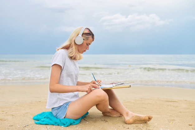 Tieners, creatieve getalenteerde jeugd, tienermeisje in koptelefoon tekenen met aquarellen mariene schets, vrouw zittend op zee strand, prachtige water landschap-achtergrond