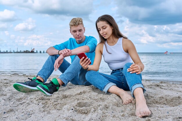 Tienervrienden zitten samen op het strand te praten en kijken naar het scherm van hun smartphone.