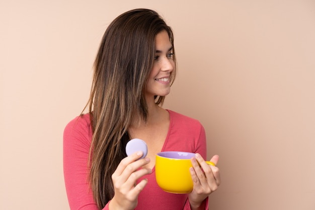 Tienervrouw over geïsoleerde muur die kleurrijke franse macarons en een kop van melk houden