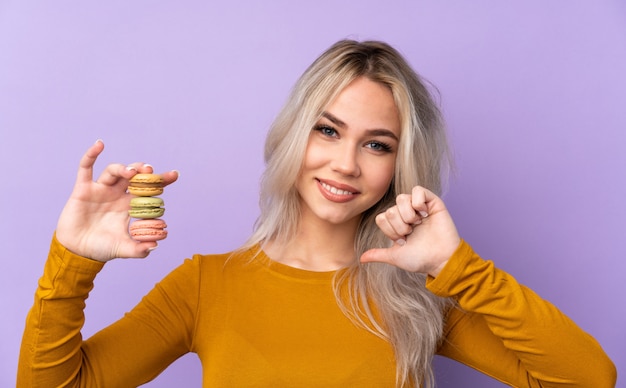 Tienervrouw over geïsoleerde purpere muur die kleurrijke Franse macarons houden en trots
