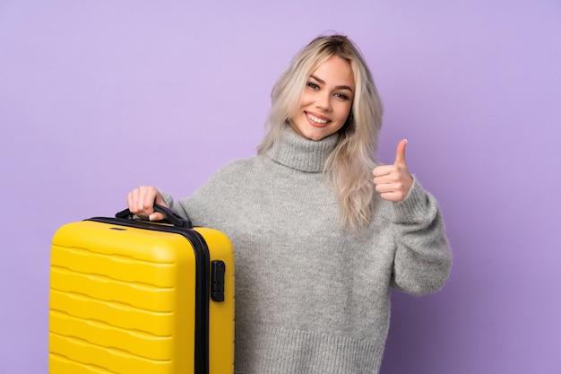 Tienervrouw over geïsoleerde purpere muur in vakantie met reiskoffer en met omhoog duim