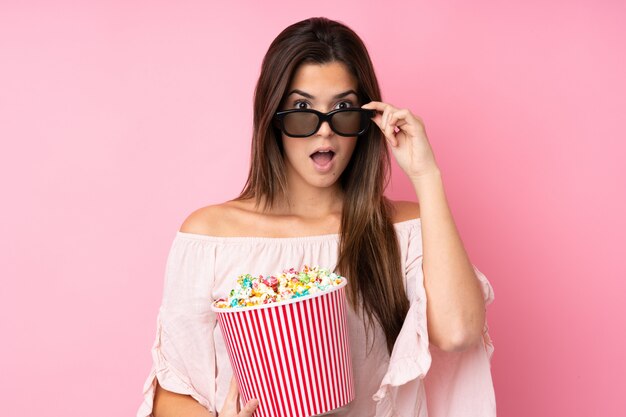 Tienervrouw over geïsoleerde roze muur die met 3d glazen wordt verrast en een grote emmer popcorns houdt