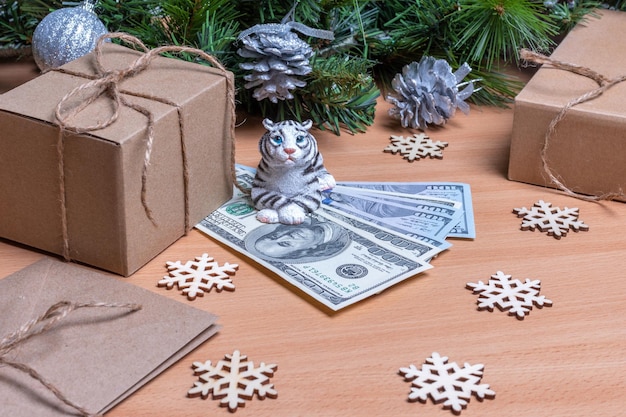The tiger is the symbol of the Chinese new year 2022. Toy tiger holding cash dollars on a wooden background with gift boxes, snowflakes and fir branches with cones. Success and wealth concept