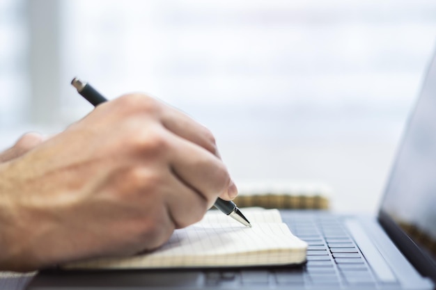 Foto colpo stretto della mano di un uomo che scrive in un blocco note su uno sfondo elegante per laptop sfocato