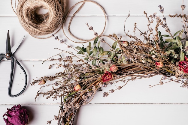 Photo tightly woven handmade dry herbal wreath from twigs skein of thread and scissors nearby white planking table on background florist crafting tutorial tradition festive decor as present top view