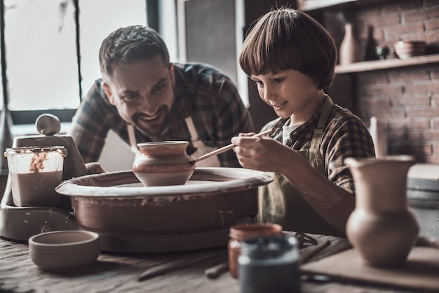 Tijd om er iets moois van te maken. Kleine jongen tekenen op keramische pot bij de pottenbakkersles terwijl man in schort dicht bij hem staat