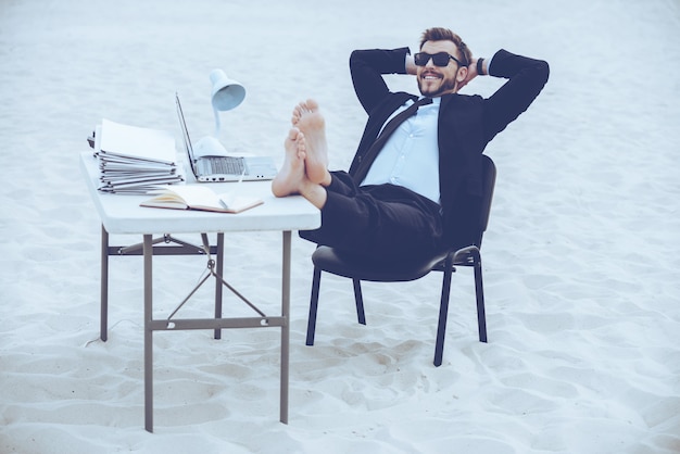 Tijd om te ontspannen. Knappe jonge man in formalwear en zonnebril hand in hand achter het hoofd en met zijn voeten op de tafel staande op zand