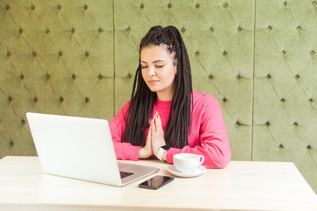 Tijd om te ontspannen. Succesvolle, aantrekkelijke jonge meisjesfreelancer met zwart dreadlocks-kapsel zit in café en rust uit, hand in hand zoals meditatief, heb emotioneel plezier. binnen