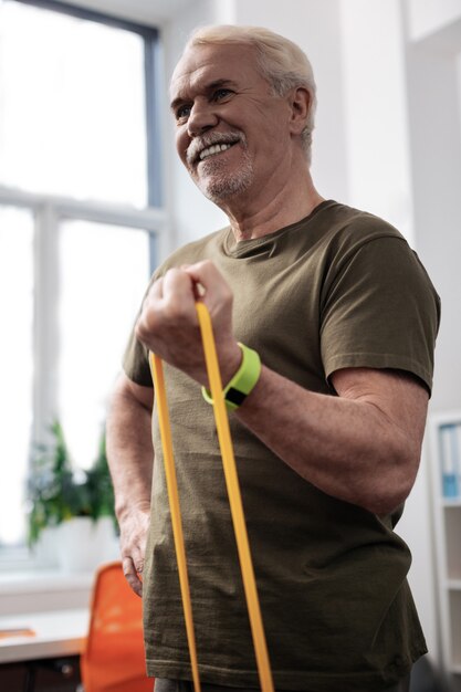 Tijd om te trainen. Opgetogen vrolijke man die in de oefenruimte staat tijdens het trainen met een rubberen band