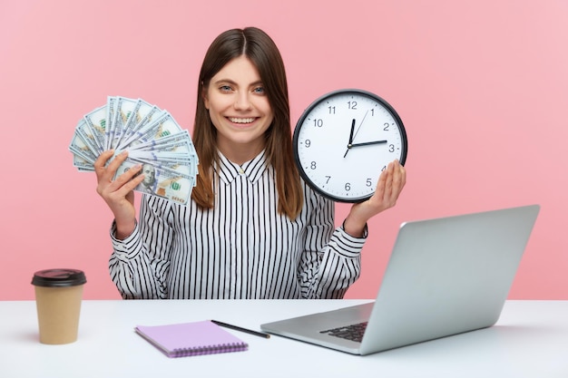 Tijd om winst te maken Positieve gelukkig zakenvrouw met Wandklok en dollar biljetten kijken camera met brede glimlach zittend op de werkplek Indoor studio opname geïsoleerd op roze achtergrond