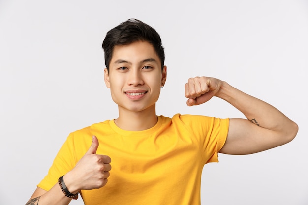 Tijd pomp het op. Aantrekkelijke jonge Aziatische man in geel t-shirt met spieren en duim-omhoog, glimlachend, moedigen aan fysieke oefeningen te doen, samen gym te komen, sterke biceps te willen