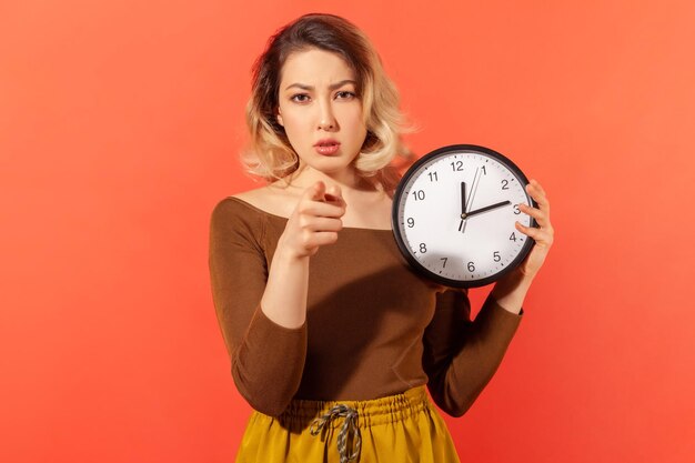 Time to act serious woman holding big wall clock pointing finger at you motivation warning about deadline indoor studio shot isolated on orange background