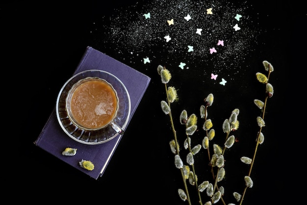 Time for a cup of hot coffee a cup of coffee stands on a book next to it is a bouquet of flowering willow two heartshaped sugar cubes a dark background top view