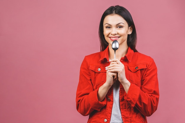 Time for dinner! Portrait of young beautiful woman wearing red casual shirt, holding spoon isolated over pink background.