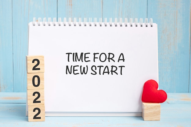 青い木製のテーブルの背景に赤いハートの形の装飾が施された新しいスタートワードと2022キューブの時間。新年newyou、目標、決意、健康、愛と幸せなバレンタインデーのコンセプト