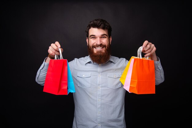 쇼핑할 시간! 수염을 기른 젊은 남자가 검은색 배경에서 쇼핑하기 위해 5개의 다채로운 종이 가방을 들고 있습니다.