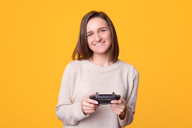 Time for fun. Portrait of smiling young woman using controller standing over yellow background