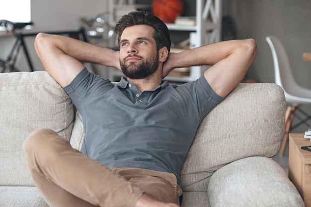 Time to gather all ideas in his head. Handsome young man keeping hands behind his head and looking thoughtful while sitting on the couch at home