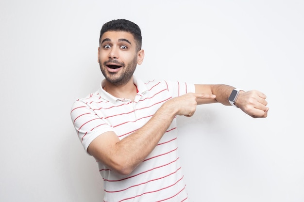 Time to go. Portrait of amazed funny bearded young man in striped t-shirt standing, pointing at his smart watch, looking at camera with surprised face. indoor studio shot, isolated on white background